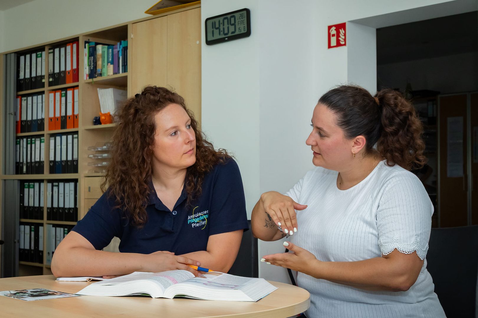Zwei Frauen sitzen an einem Tisch, umgeben von Büchern und Unterlagen, und diskutieren angeregt.
